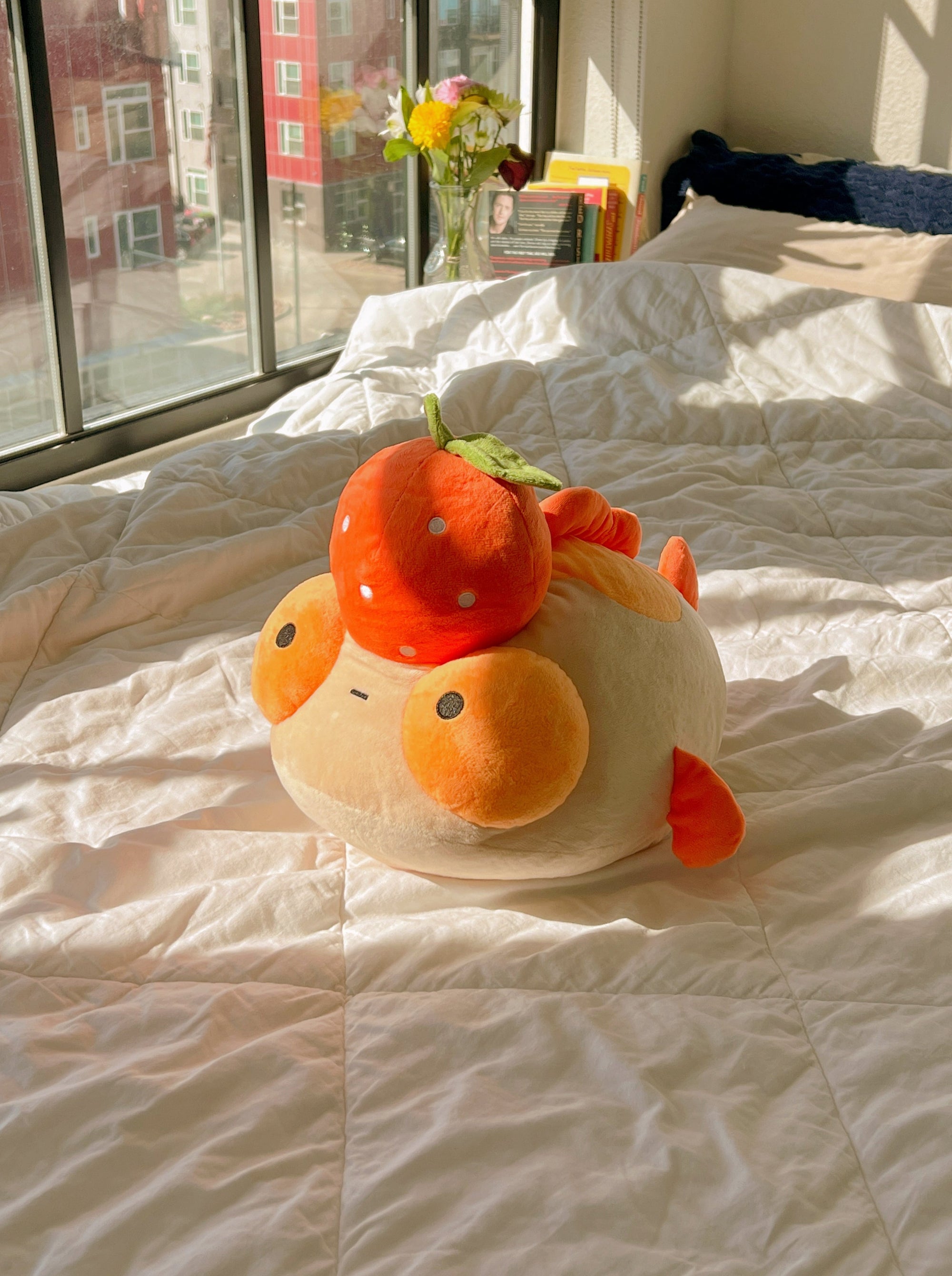 An orange plush goldfish with a tangerine on it's head, sitting on a bed with a white quilt.
