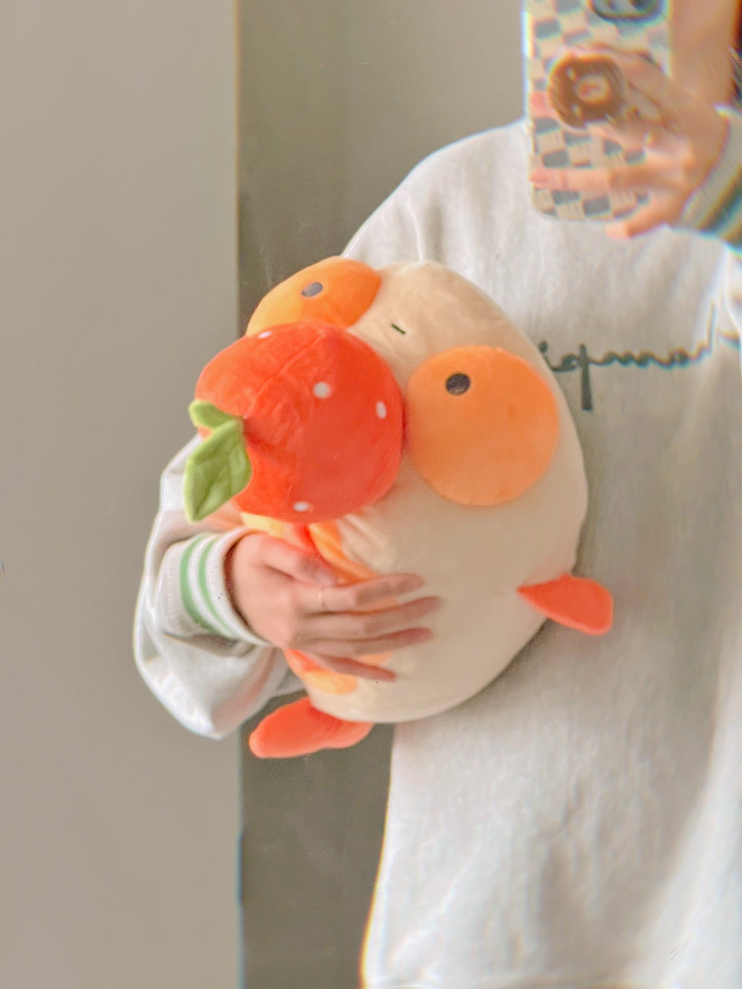 An orange plush goldfish with a tangerine on it's head, being held under an arm in a mirror selfie. 