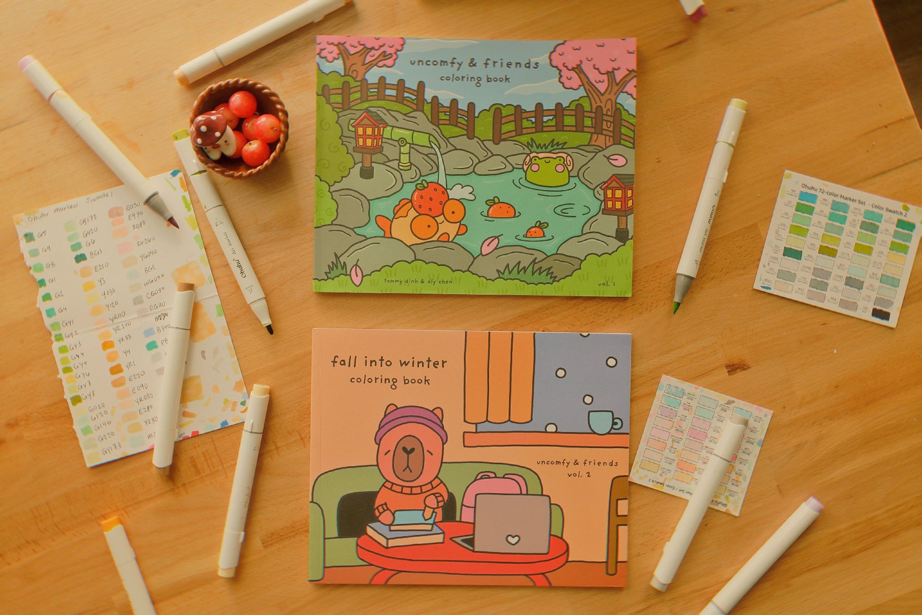 Two coloring books on a wooden table with markers and a small bowl of apples.