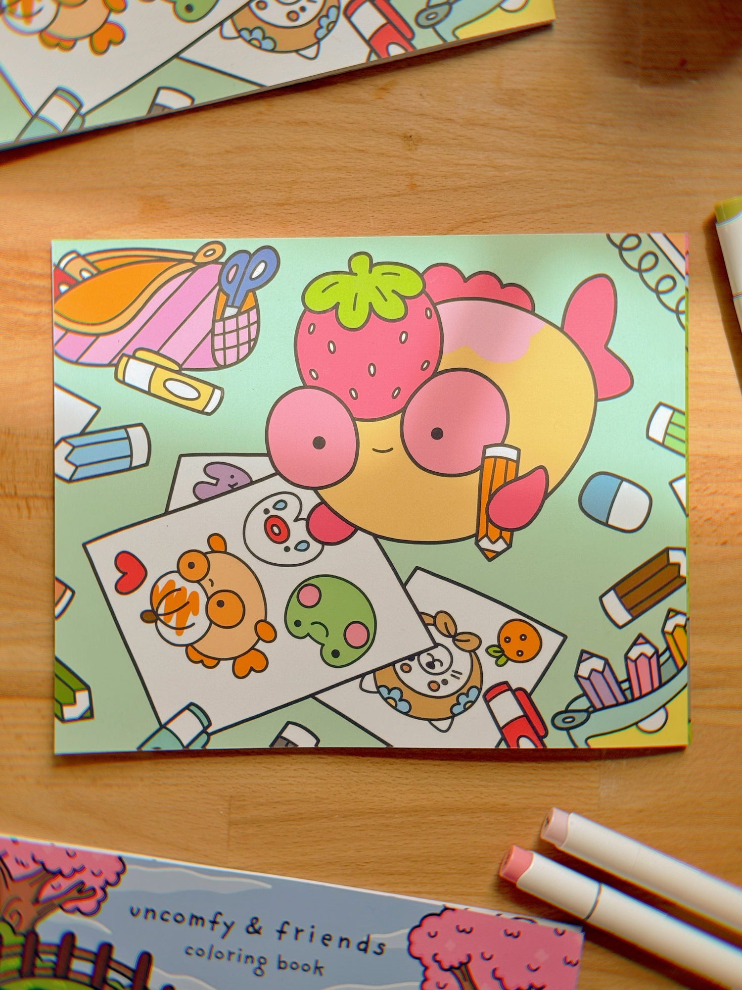 The back of the coloring book feauring an image of Strawberry Maxine coloring in Pumpkin Maxine, on a wooden surface with markers next to it. 